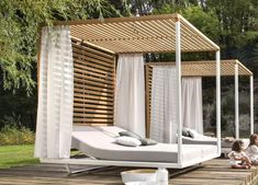 two children sitting on a wooden deck next to a bed with white sheets and drapes