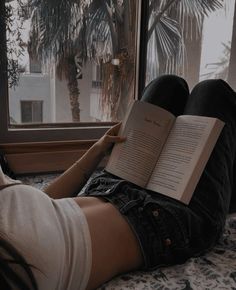 a woman laying on her stomach reading a book in front of a window with palm trees outside