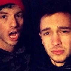 two young men posing for the camera in front of a dark background, one is wearing a red beanie