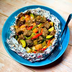 a blue plate topped with meat and veggies covered in tin foil next to a fork