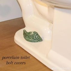 a green fish figurine sitting on top of a white toilet seat next to a wooden floor