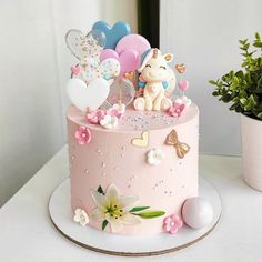 a pink birthday cake decorated with balloons, flowers and other decorations on a white table next to a potted plant