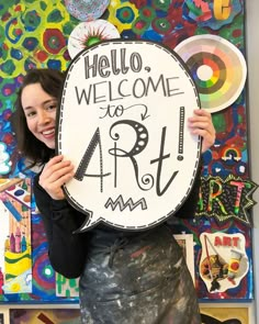 a woman holding up a sign that says hello, welcome to the art room in front of her