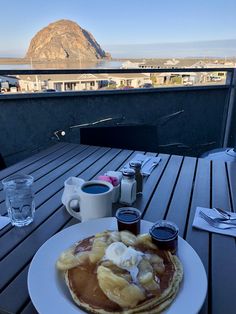 pancakes with butter and syrup on a white plate sitting on a wooden table overlooking the ocean