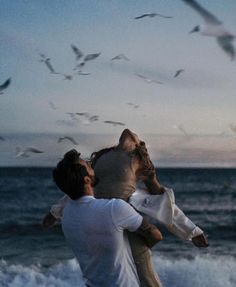 two people on the beach with seagulls flying in the sky above them and one person holding onto another man's shoulders