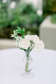 white flowers are in a clear vase on a round table with greenery behind it