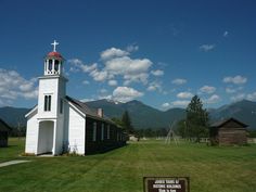 a small white church with a steeple in the grass and mountains in the background