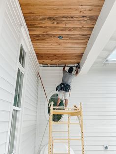 a man standing on a ladder working on a wood paneled ceiling above a white house