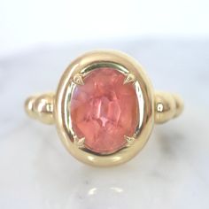 This one-of-a-kind statement ring features a vibrant orange-pink tourmaline as its centerpiece. Set in 14K yellow gold, the tourmaline is encircled by a chunky golden bezel a unique bubble-like design on the side of the band. This custom-designed ring is perfect for adding a pop of color to any outfit and making a bold fashion statement. The orange-pink tourmaline is a rare and unique gemstone and the yellow gold band gives the ring a warm and elegant look. The ring is unique and has a special d Pink Tourmaline Rings, Opal And Pink Tourmaline Ring, Orange Engagement Ring, Pink Tourmaline Rings As Gift, Oval Pink Tourmaline Ruby Ring, Pink Tourmaline Yellow Gold Ring, Pink Tourmaline Engagement Ring, Pink Engagement Ring, Bezel Engagement Ring