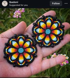 two handmade beaded earrings in the palm of someone's hand, with flowers behind them