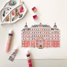 some watercolors and markers are on the table next to a drawing of a pink building