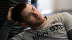 a man sitting down with his hand on his head and looking at the camera while wearing a gray shirt