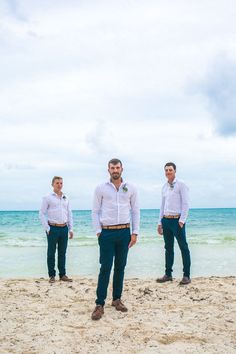 three men are standing on the beach in front of the ocean and one man is wearing a white shirt and blue pants