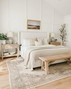 a bedroom with white walls and wood flooring has a large bed, two nightstands, and a bench in front of the bed