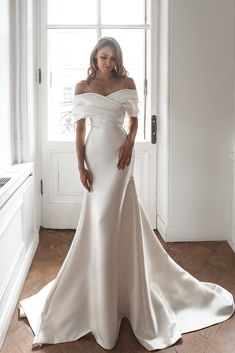 a woman standing in front of a window wearing a wedding dress with off the shoulder sleeves