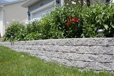 a stone retaining wall in front of a house with flowers growing on the side and bushes behind it