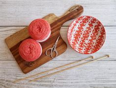 two skeins of yarn next to a bowl and pair of knitting needles on a cutting board