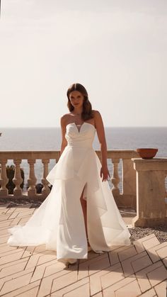 a woman in a white wedding dress standing on a balcony next to the ocean and looking at the camera