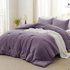 a bed with purple comforter and pillows in a white room next to a window