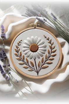 a white and brown flower is on a table next to some lavenders, with a cross - stitch pattern in the middle