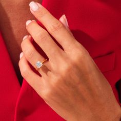 a woman's hand wearing a gold ring with a diamond in the middle and a red dress behind her