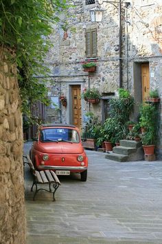 an old red car is parked in front of a stone building with potted plants