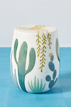 a white stool with green and gold painted cactus designs on it, sitting on a blue surface