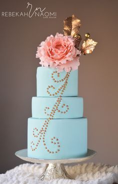 a three tiered blue cake with pink flowers and gold leaves on top, sitting on a table