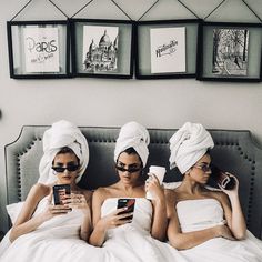 three women laying in bed with towels wrapped around their heads looking at cell phones and drinking coffee