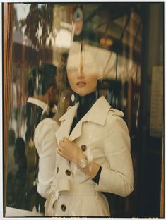 a woman in a white trench coat looking through a window with her hands on her hips