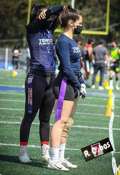 two female football players standing on the sidelines with their hands behind their backs, wearing face masks