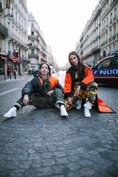 two young women sitting on the ground in front of a police car, one wearing an orange and black jacket