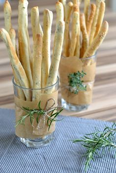 some bread sticks are in a small glass cup on a table with rosemary sprigs