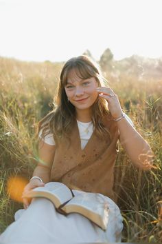 a young woman sitting in the grass reading a book and talking on her cell phone