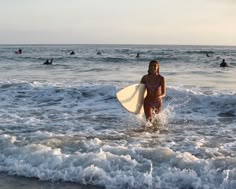 a woman walking into the ocean with a surfboard in her hand and people swimming in the water behind her