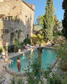 an outdoor swimming pool surrounded by trees and people in the water, with stone buildings behind it