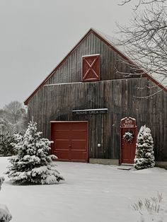 a barn with two red doors and snow covered trees in the foreground on a snowy day