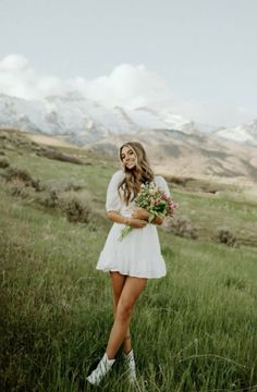 a woman in a white dress is standing in the grass with flowers and mountains behind her