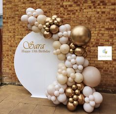 a large balloon arch with gold and white balloons in front of a brick wall that says ooay happy 13th birthday