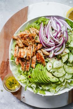 a salad with chicken, cucumbers and onions on a white plate next to a lemon wedge