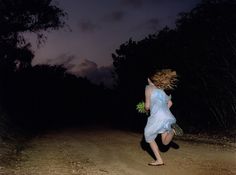 a woman in a blue dress running down a dirt road at night with her hair blowing in the wind