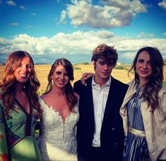 three women and a man posing for a photo in front of a wheat field with clouds