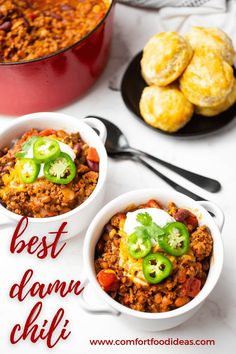 two white bowls filled with chili and cheese next to muffins on a table