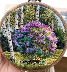 a person holding up a small embroidery art piece with trees and flowers painted on it