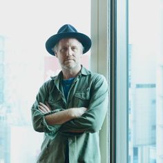 a man wearing a hat standing in front of a window with his arms crossed and looking at the camera