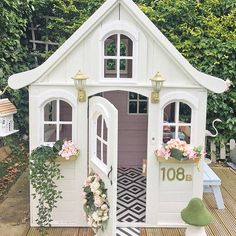 a white doll house with windows and flowers on the front door is sitting on a wooden deck