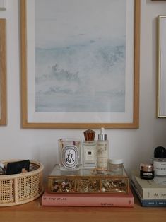 two framed pictures hang on the wall above a table with perfumes and books in front of it