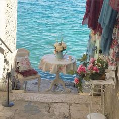 there is a table with flowers on it next to some chairs and a chair near the water