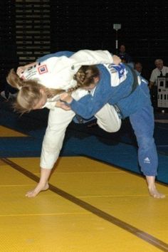 two people in blue and white uniforms are doing karate moves on a yellow mat while others watch