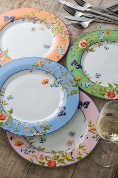 four colorful plates with flowers on them next to wine glasses and silverware set on a wooden table